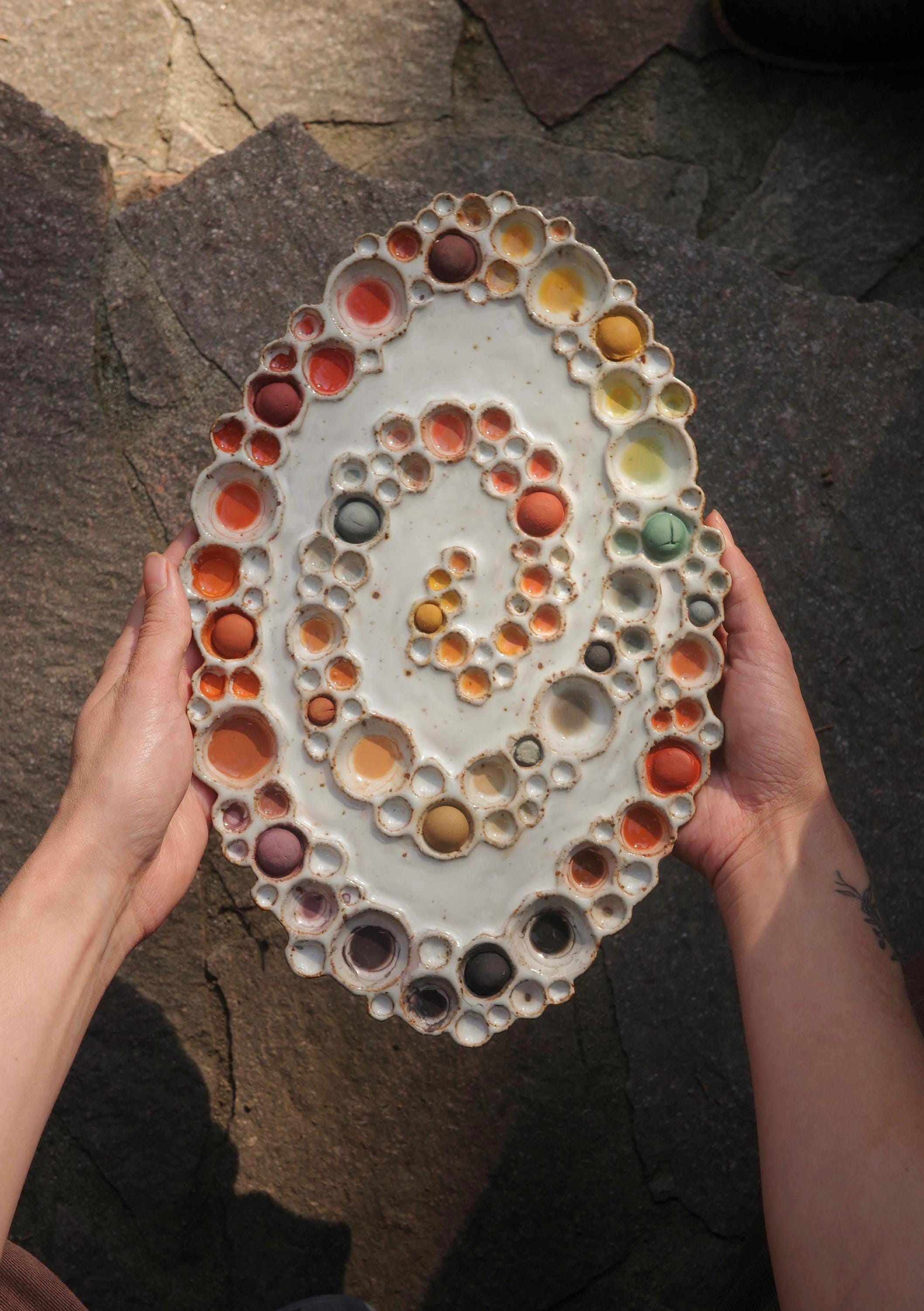 Two hands holding ceramic spiral bubble palette with rainbow paints and a hand holding a paintbrush against gray stone background 