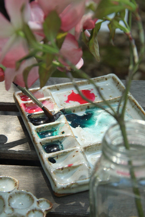 Ceramic rectangular paint palette with paint swatches on a wooden surface with flowers.