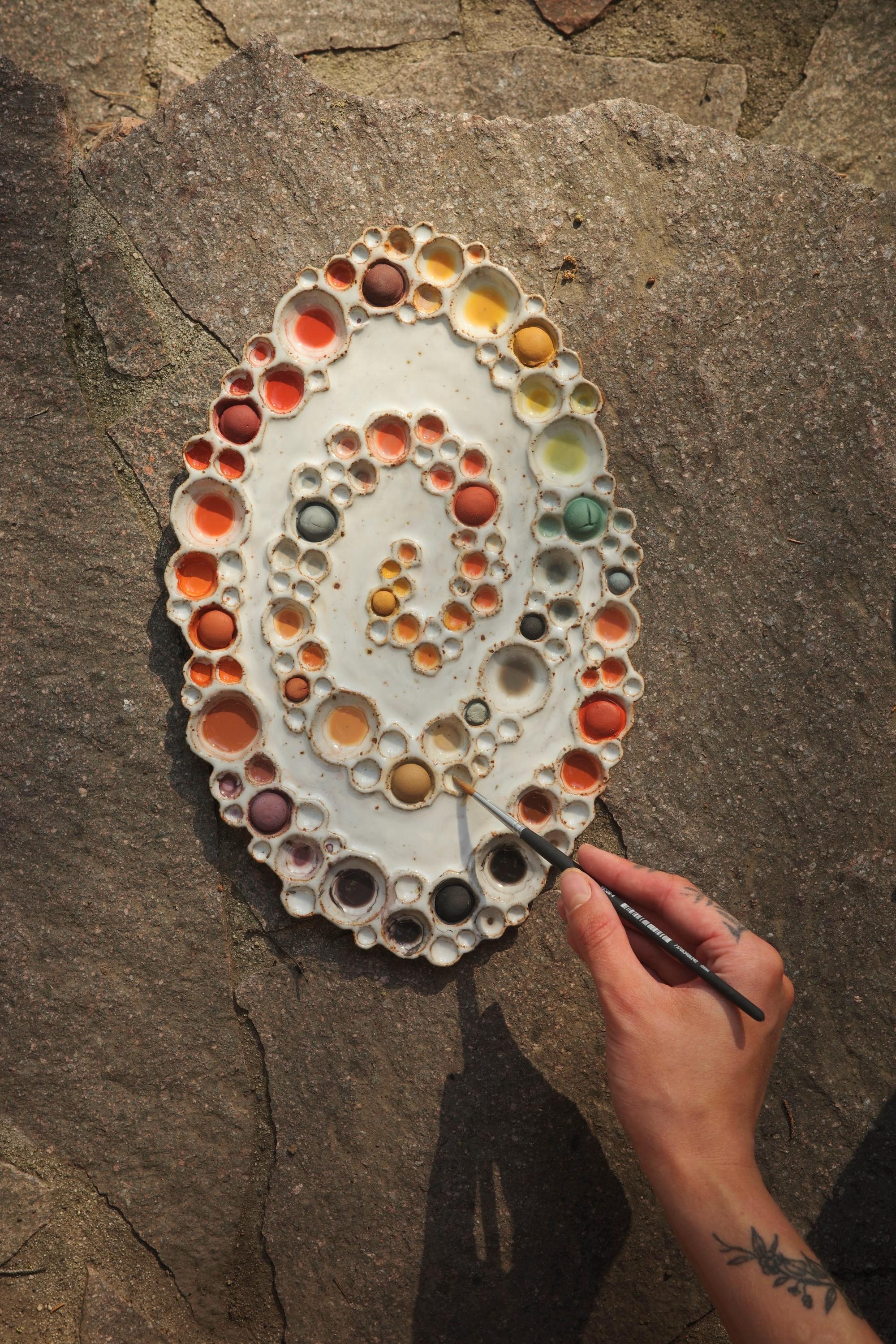 Ceramic spiral bubble palette with rainbow paints and a hand holding a paintbrush against gray stone background 