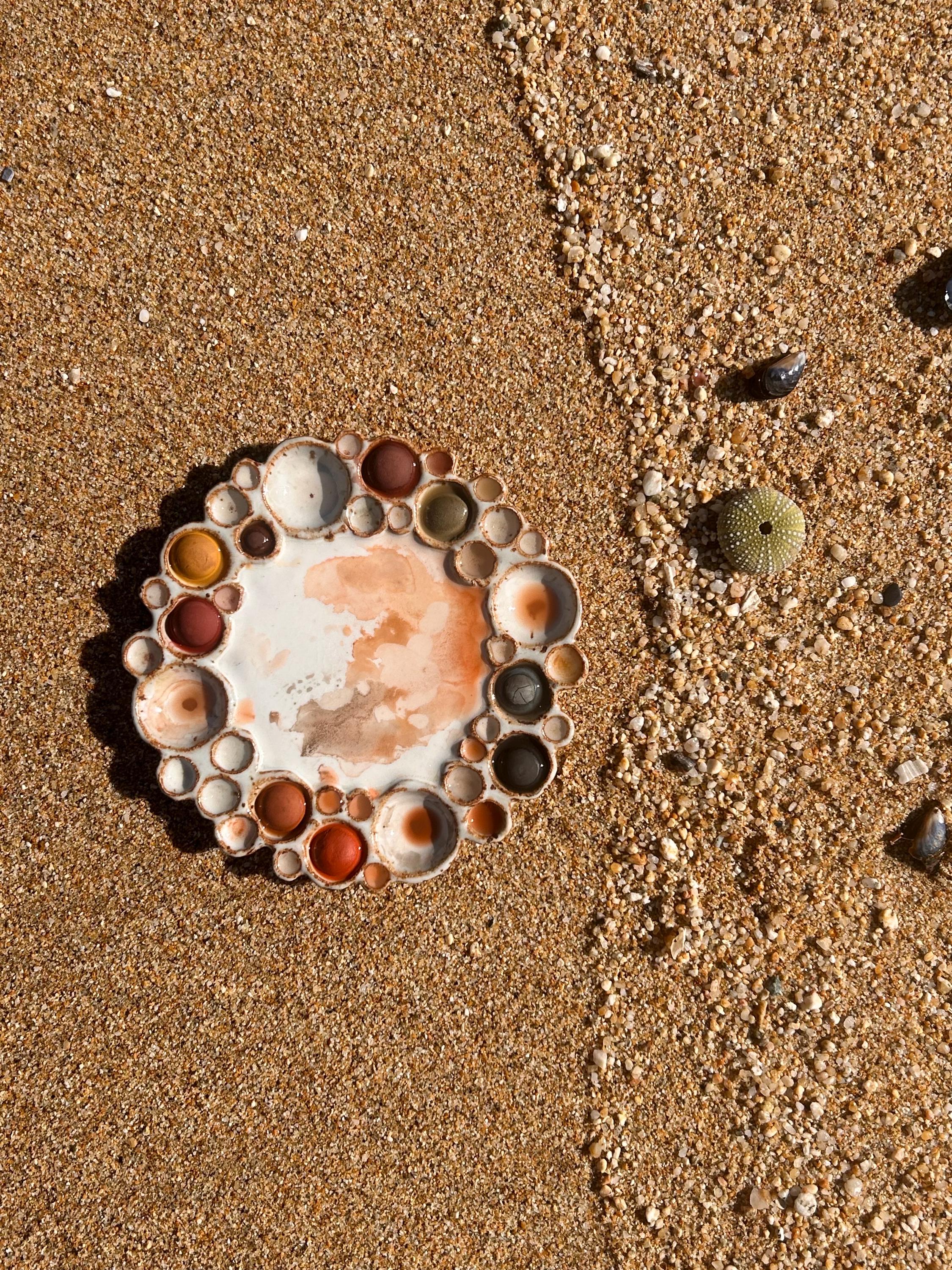 Round watercolor palette with wells with paint inside them on a sandy beach with shells 
